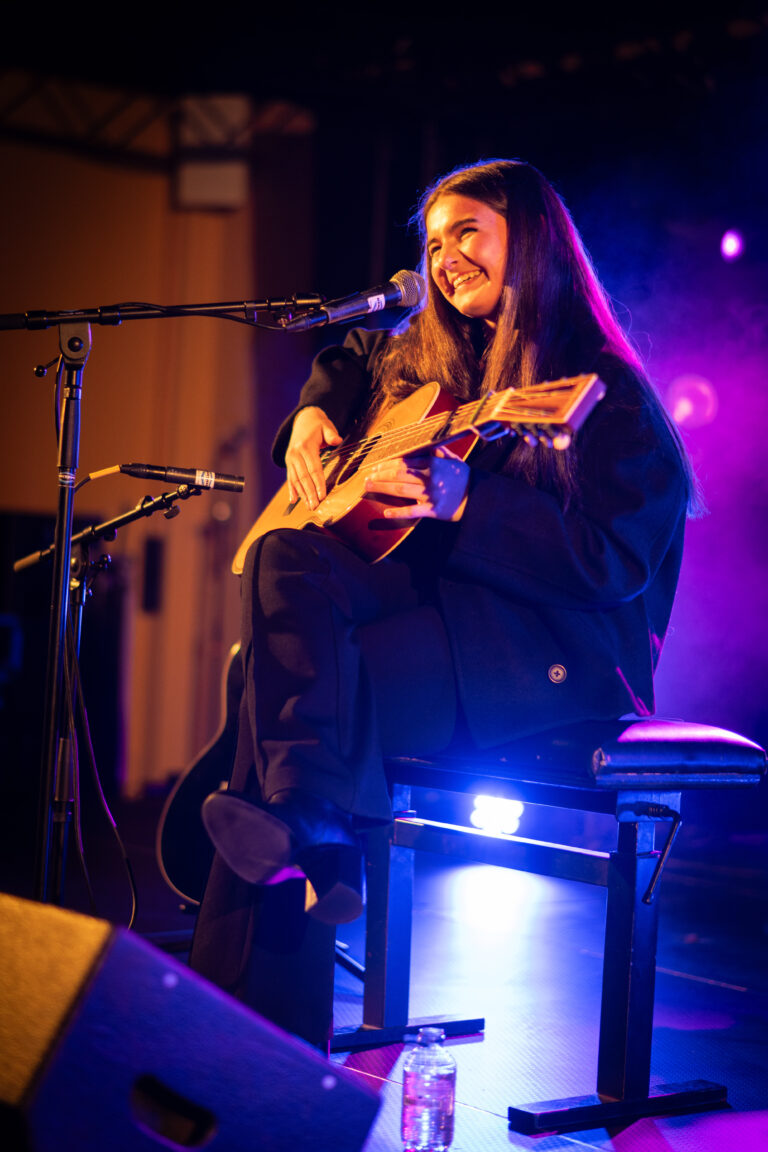 Een vrouwelijke muzikant zit op een stoel met een gitaar in de hand, terwijl ze een optreden geeft onder kleurrijke lichten.