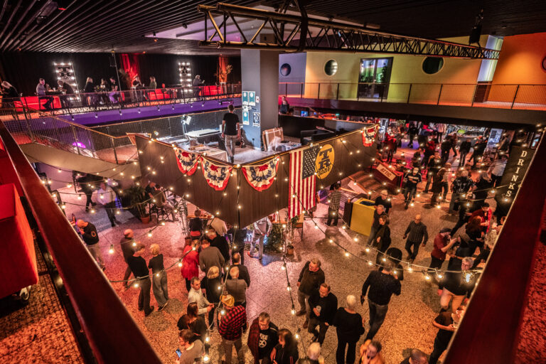 Een drukke evenementenlocatie met mensen die zich verzameld hebben in een sfeervol verlichte ruimte, versierd met een Amerikaanse vlag en feestelijke decoraties.