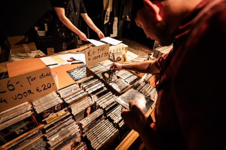 Een man kijkt naar cd's op een verkooptafel, terwijl iemand anders op de achtergrond aanwezig is.