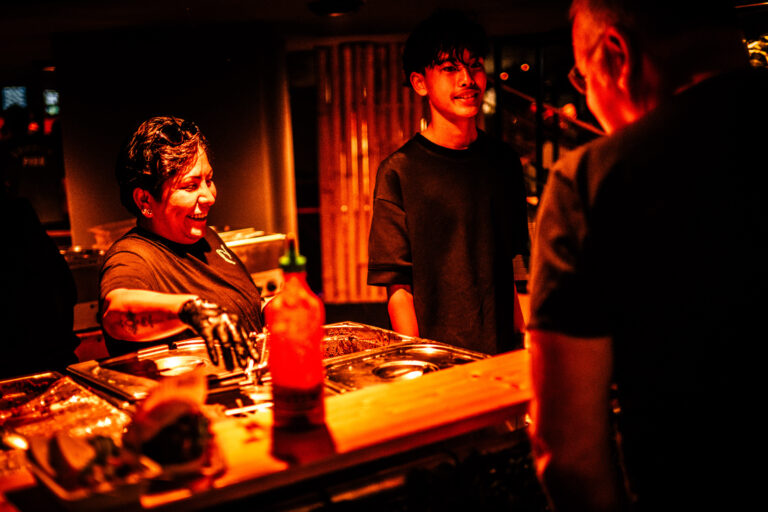 Een vrouw met een glimlach staat achter een bar terwijl ze met twee mensen praat in een sfeervol verlichte ruimte.