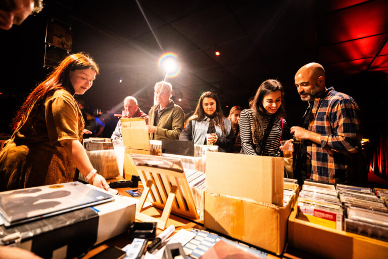 Een groep mensen kijkt naar en sorteert platen en cd's op een levendig evenement in een donkere ruimte.