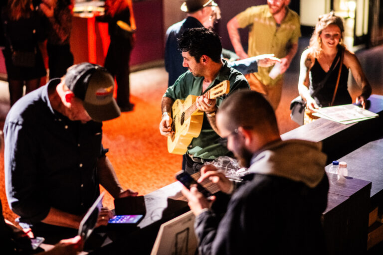 Een muzikant op een podium speelt gitaar terwijl bezoekers interactie hebben aan de bar.