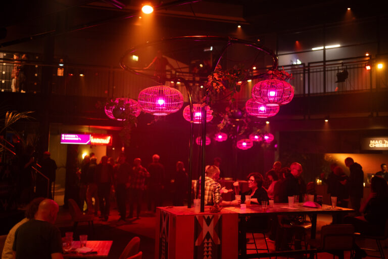 Een sfeervolle binnenruimte met mensen, verlicht door roze lampen en een bar in het midden.