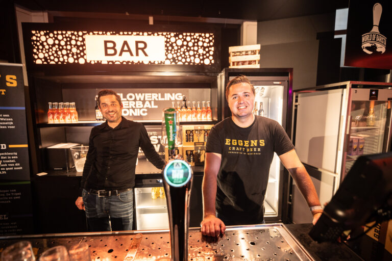 Twee barmannen staan achter een bar met een tap en koelkasten, terwijl een lichtgevend 