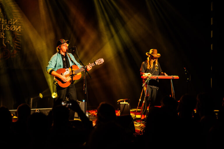 Twee muzikanten geven een live optreden op het podium, met een gitaar en een steelgitaar, omringd door sfeervolle lichten.