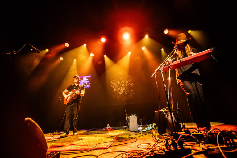Twee muzikanten optreden op een podium met kleurrijke verlichting, waarbij de ene een gitaar bespeelt en de andere toetsen speelt.