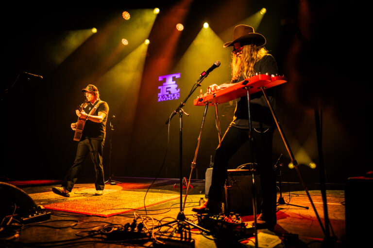 Twee muzikanten voeren op een podium met kleurrijke lichten, waarbij één gitarist speelt en de ander een keyboard bespeelt.