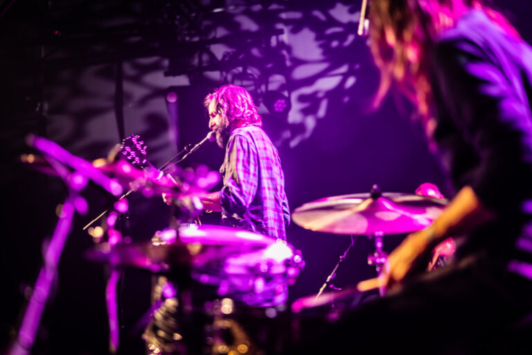 Een muzikant speelt gitaar en zingt op een podium, terwijl een andere muzikant op de drums speelt, verlicht door paarse lichten.