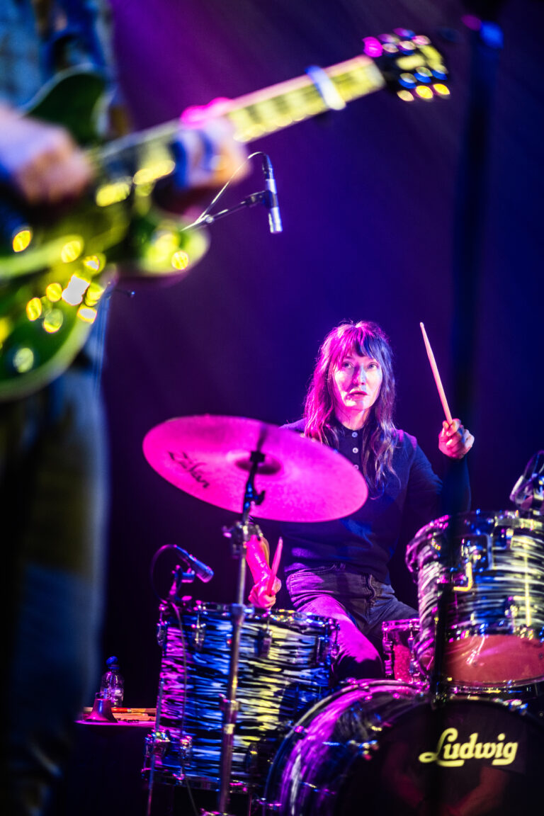 Een vrouwelijke drummer speelt op een kleurrensset tijdens een live optreden, terwijl een muzikant op een gitaar op de voorgrond staat.