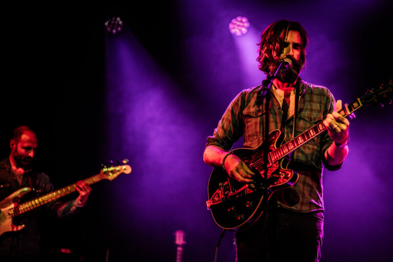 Een man speelt gitaar op een podium, terwijl een andere muzikant op de achtergrond een basgitaar bespeelt, met paarsachtige lichteffecten.