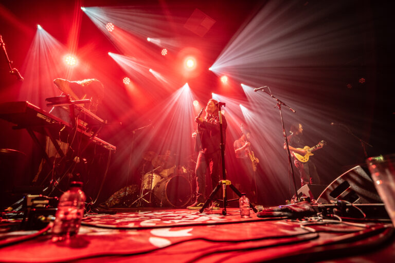 Een band speelt live op het podium met felrood licht en zichtbare muziekinstrumenten.