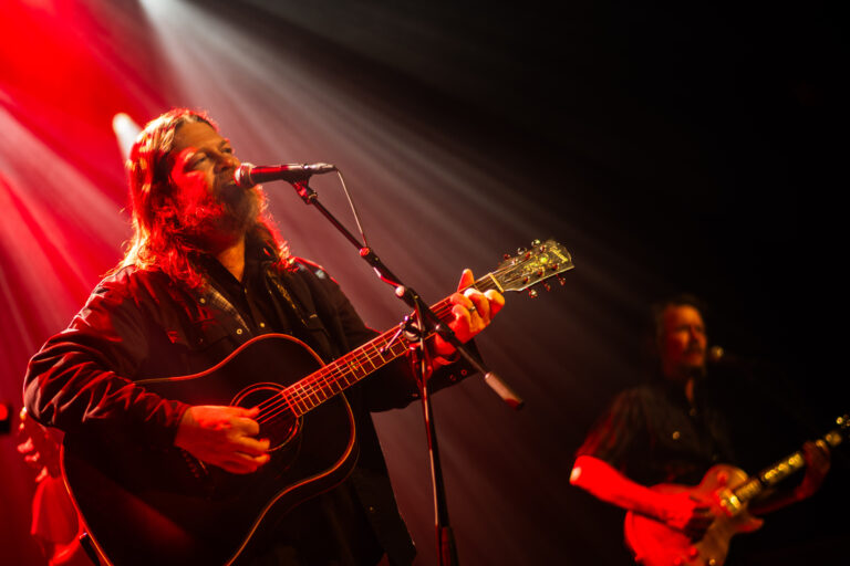 Een muzikant met een gitaar zingt op het podium terwijl een andere muzikant op de achtergrond speelt, omringd door sfeervol licht.