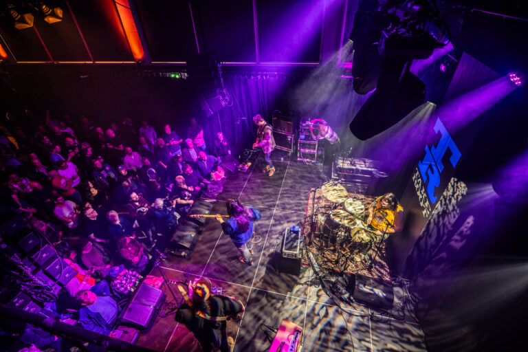 Een bovenaanzicht van een muzikale live-optreden met een band op het podium en een publiek dat enthousiast kijkt, verlicht door paarse spotlights.