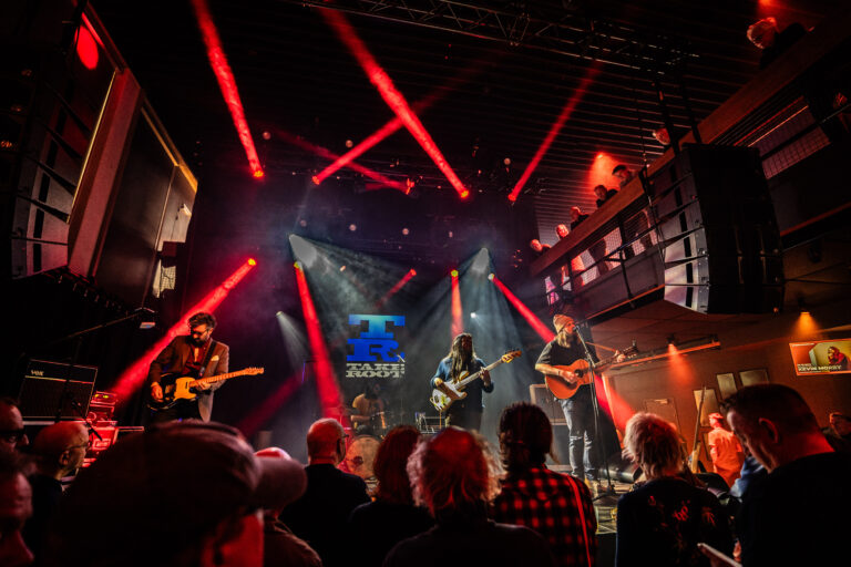 Een band speelt live op een podium, verlicht door felrode lichten, terwijl een menigte toekijkt.