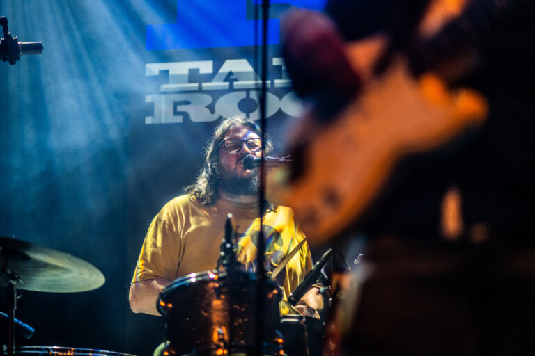 Een drummer in een geel shirt speelt op het podium, omringd door lichtstralen en schaduwen.