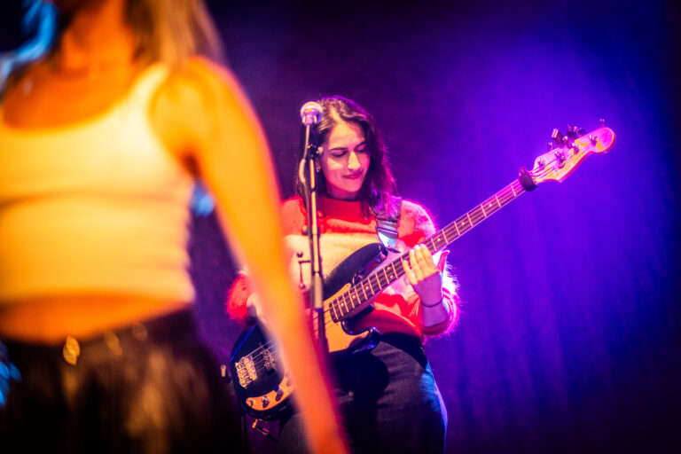 Een muzikante speelt basgitaar op het podium terwijl een andere performer in het voorgrond zichtbaar is.