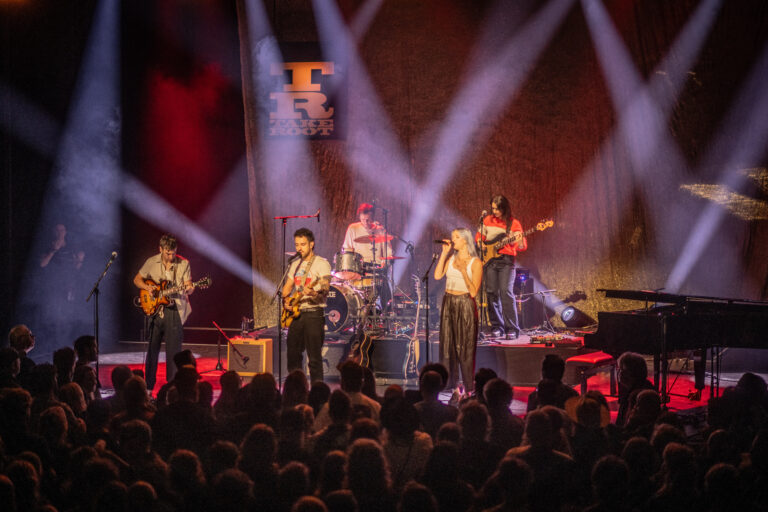 Een band speelt live op een podium voor een enthousiast publiek, omgeven door kleurrijke lichtstralen.