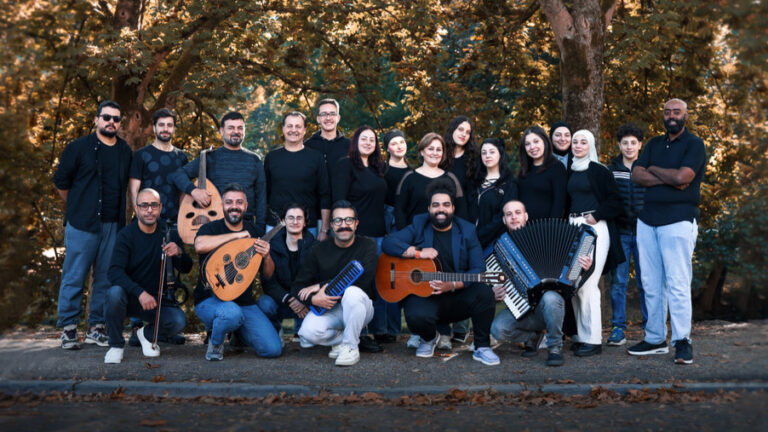 Groep mensen in zwarte kleding poseert samen buiten, met verschillende muziekinstrumenten zoals gitaren en een accordeon.