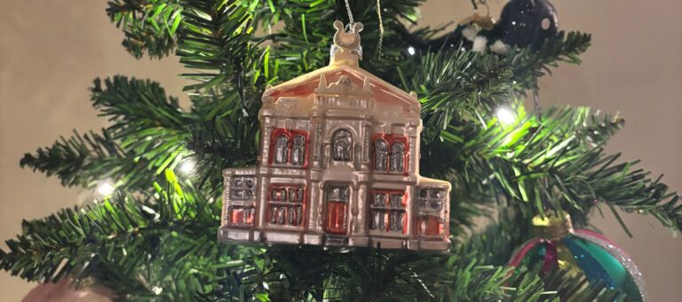 Een kerstornament in de vorm van een gebouw hangt aan een kerstboom, omringd door sierlijke lichtjes.