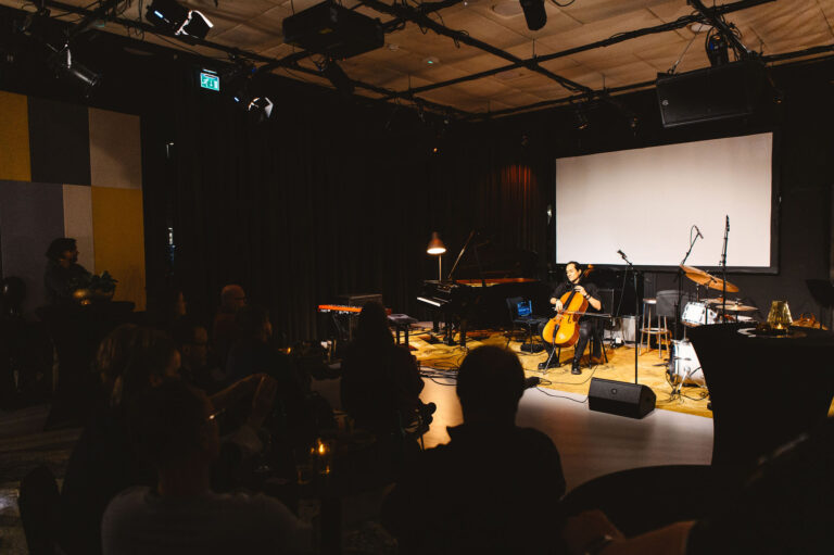 Een muzikant speelt cello op een podium voor een publiek in een intieme ruimte.