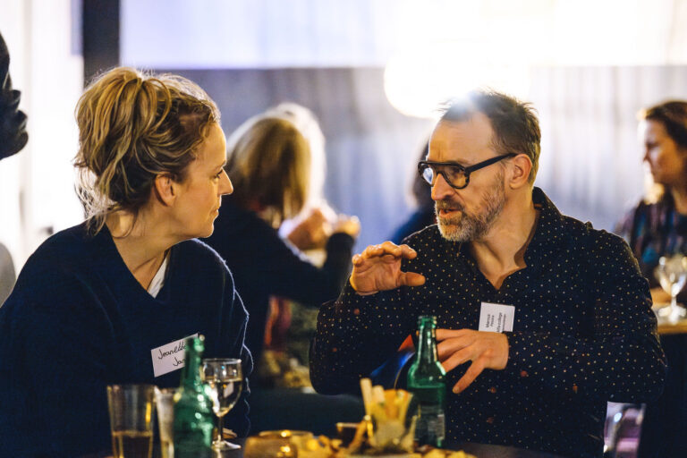 Twee mensen zitten aan een tafel en voeren een gesprek in een gezellige horecagelegenheid, met drankjes en hapjes op tafel.