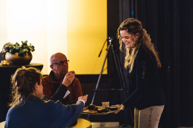 Een vrouw serveert hapjes aan twee mensen aan een tafel in een sfeervolle omgeving.