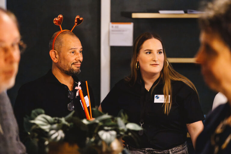 Twee panelleden zitten achter een tafel tijdens een bijeenkomst, met een plant op de voorgrond.