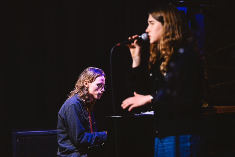 Een zangeres staat achter een microfoon op een donker podium, terwijl een tweede persoon naast haar staat.