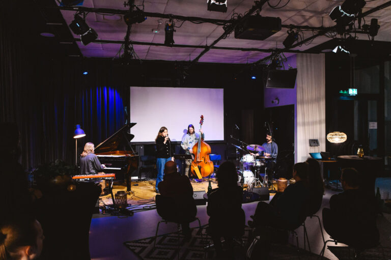 Een liveband speelt op een verlicht podium tijdens een concert, met meerdere muzikanten en een publiek.