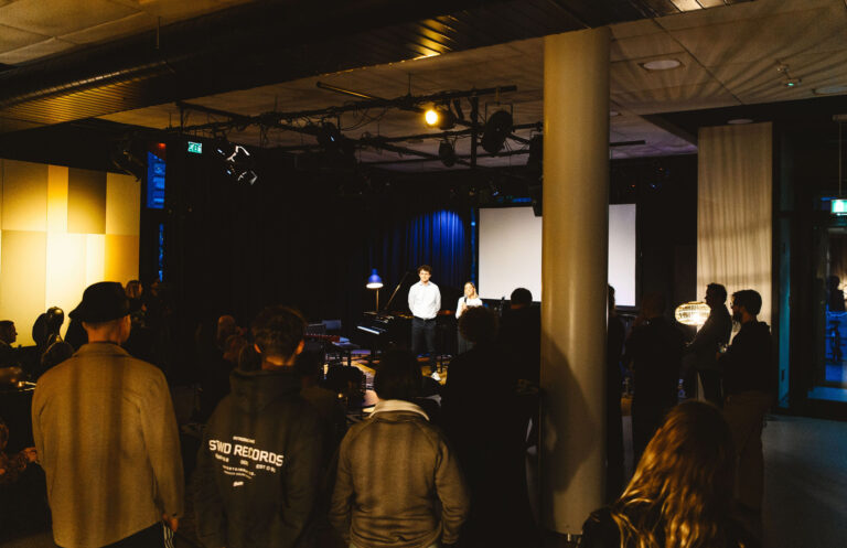 Een menigte in een donkere zaal kijkt naar een artiest die op een fel verlicht podium staat.