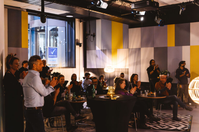Een groep mensen zit aan tafels in een moderne zaal tijdens een bijeenkomst.