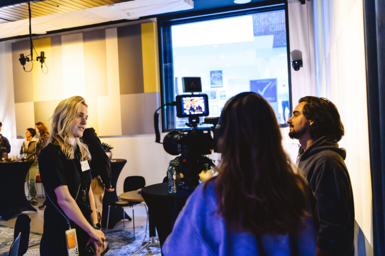 Een camera op een statief legt een groep mensen vast die aan een tafel in gesprek zijn in een studio.