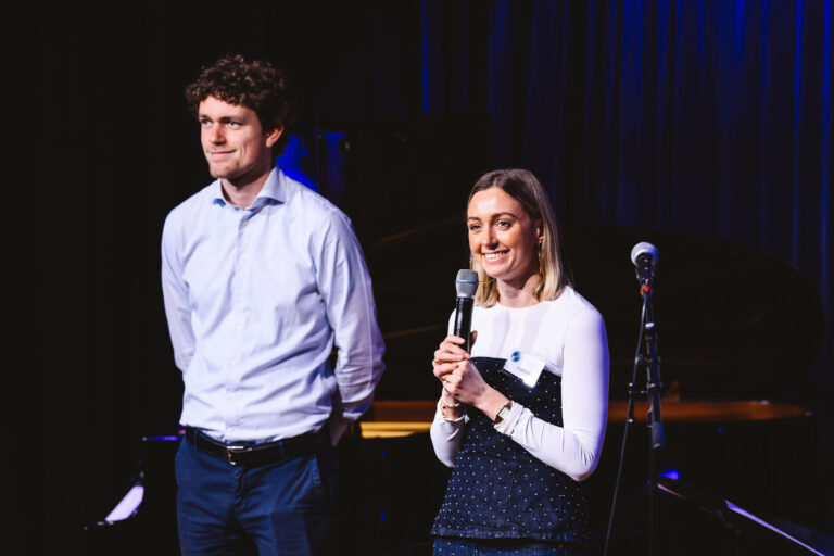 Twee presentatoren staan op een podium, de man links draagt een lichtblauw overhemd en de vrouw rechts houdt een microfoon.