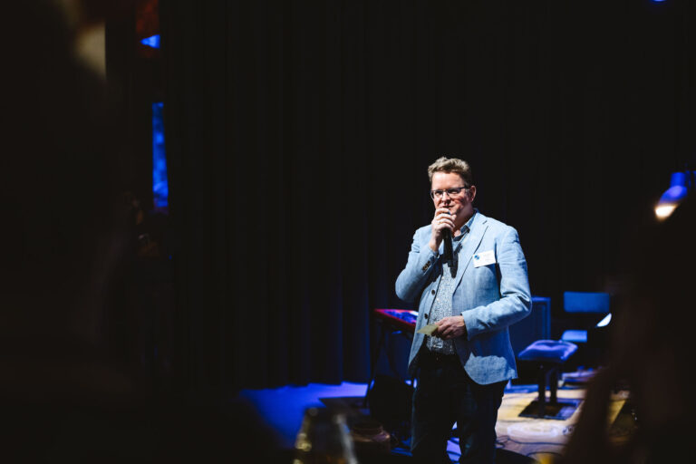 Een man in een lichtblauw jasje staat op een donker podium en houdt een microfoon vast tijdens een optreden.