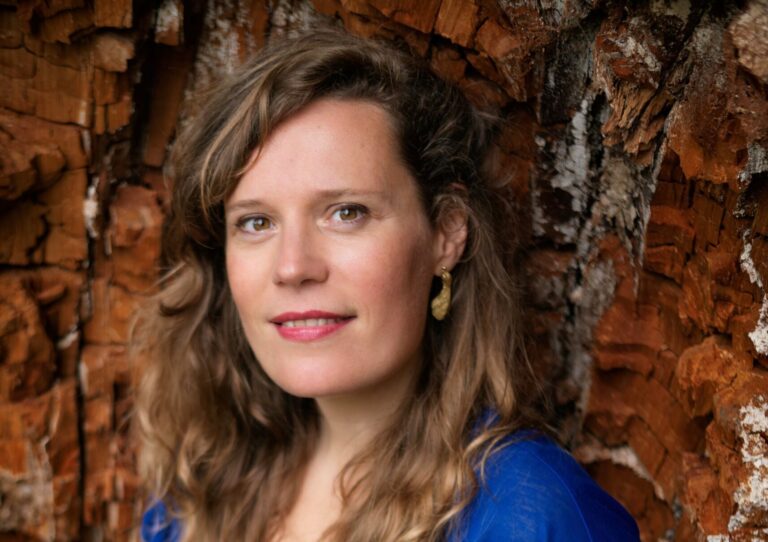 Portret van een vrouw met bruin, golvend haar tegen een bakstenen muur.