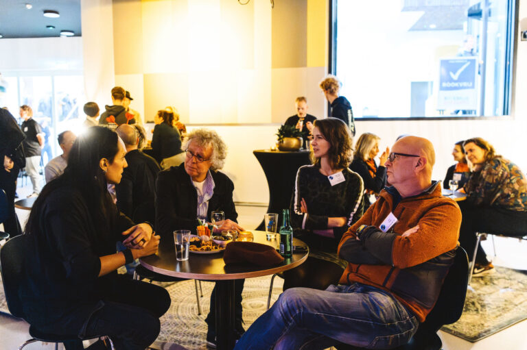 Groep mensen in gesprek rondom een tafeltje met drinken en snacks in een lichte, moderne omgeving.