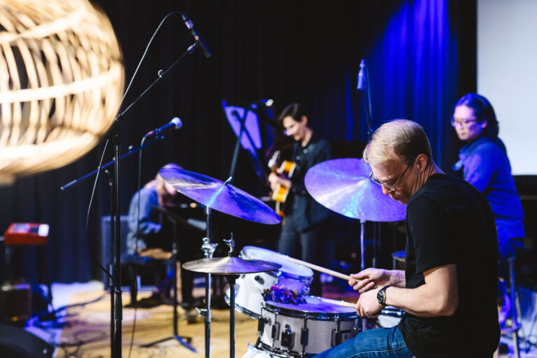 Een drummer speelt op een drumstel op een podium, met andere muzikanten op de achtergrond.