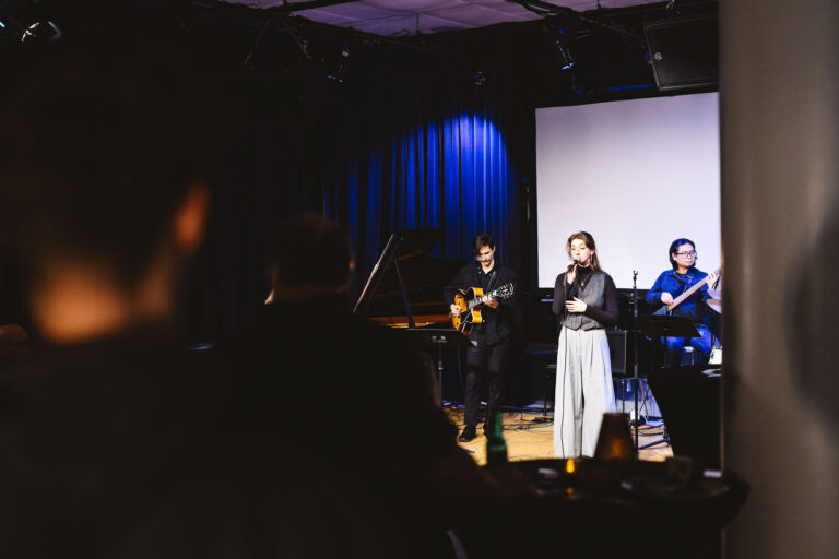 Een muzikale uitvoering met een zangeres en muzikanten op het podium, omringd door een donkere zaal.