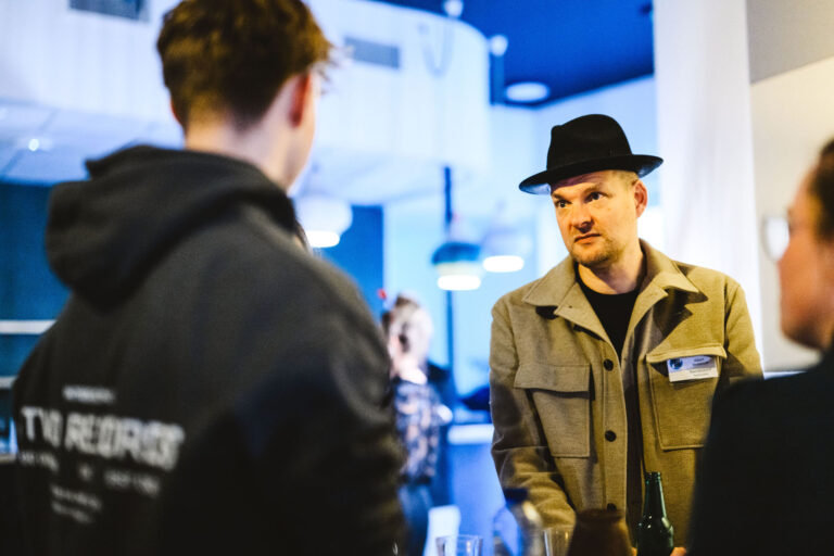 Een man met hoed en jas staat in gesprek met een andere persoon in een ruimte met blauwe verlichting.