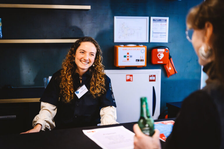 Een glimlachende vrouw met krullend haar en een naamkaartje staat achter een balie met documenten op tafel.