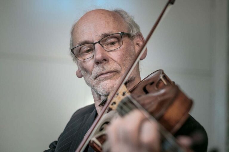 Een oudere man met een bril speelt viool met een concentrerende blik op zijn gezicht.