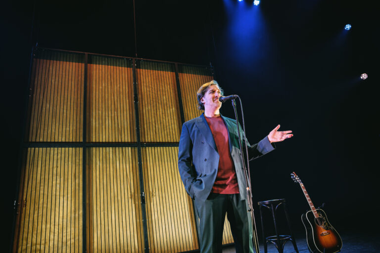 Een spreker met een microfoon staat op een podium, met een gitaar en een houten achterwand in het achtergrondlicht.