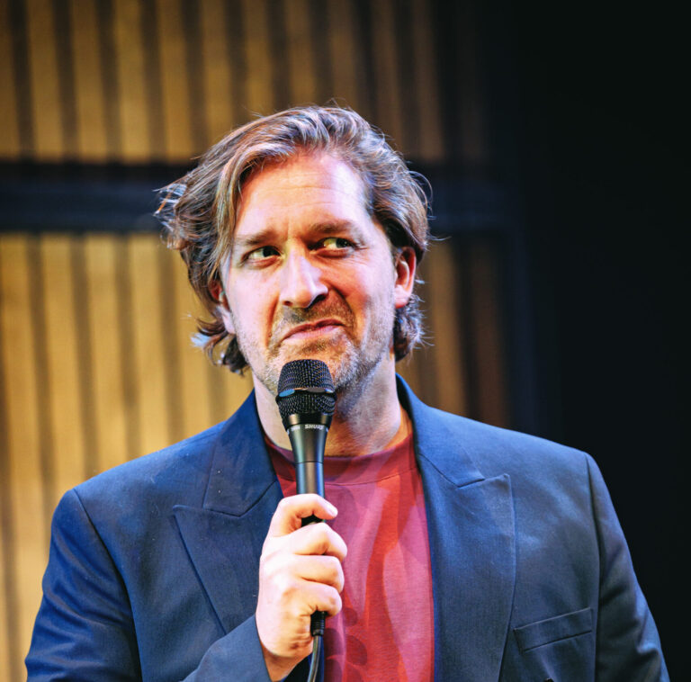 Een man met halflang haar houdt een microfoon vast terwijl hij op het podium staat.