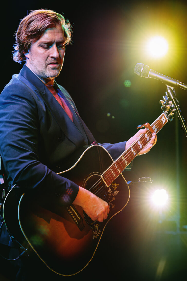 Een man in een donker pak speelt gitaar op het podium, omringd door sfeervolle verlichting.