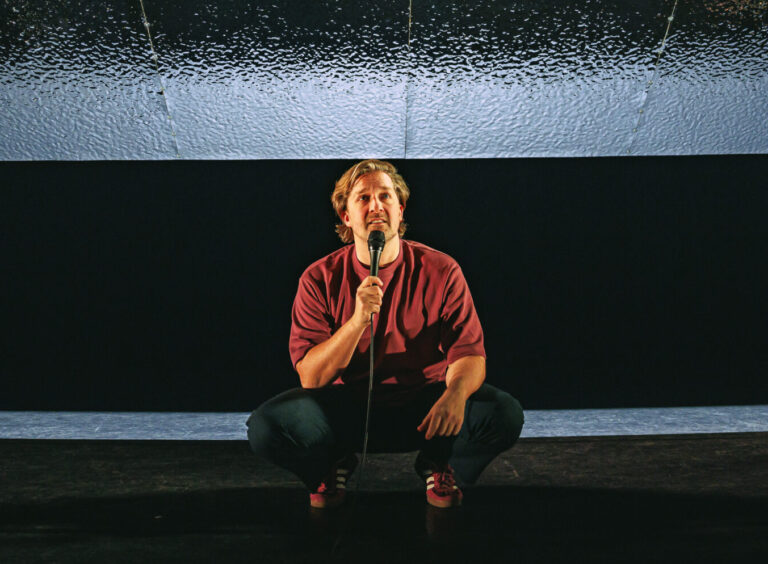 Een persoon staat in een squatpositie op een podium met een microfoon, tegen een donkere achtergrond.