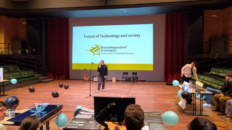 Een spreker staat op een podium in een auditorium, met een grote presentatie op het scherm achter zich.