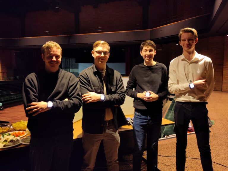 Vier mannen staan met elkaar in een ruimte, met hun armen over elkaar en een tafel met snacks op de achtergrond.