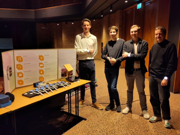 Een groep van vier mensen staat naast een tafel met presentatiemateriaal en technologie, waaronder gadgets en een groot bord met informatie.