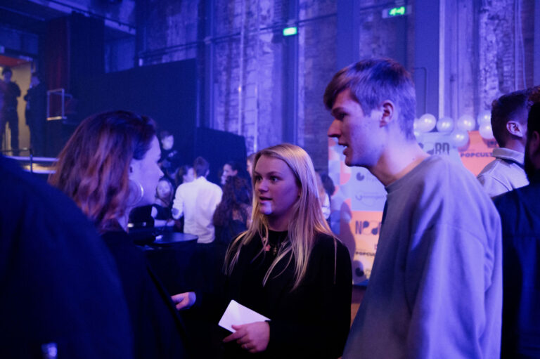 Drie mensen staan in gesprek in een sfeervolle ruimte tijdens een evenement met kleurrijke decoratie op de achtergrond.