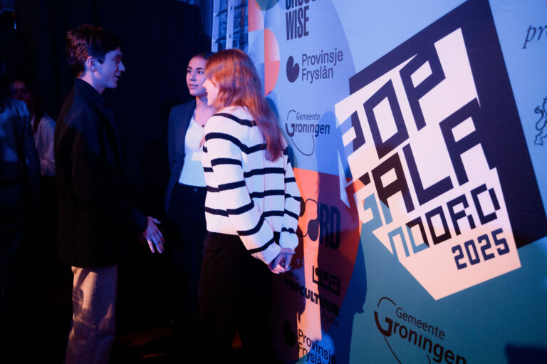 Drie mensen staan in gesprek bij een kleurrijke muur met het logo van Pop Gala Noord 2025.